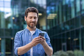 Smiling businessman with beard using smartphone outside modern office