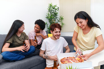 Multicultural group of friends sitting together on a sofa, enjoying pizza and beers, celebrating a birthday in a warm home setting