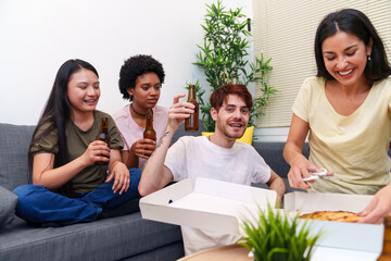 Diverse friends enjoy a casual birthday party at home, eating pizza and drinking beers together on a comfortable sofa.