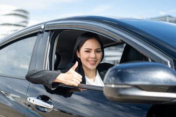 Young beautiful asian business women in suit getting new car showing thumbs up. She very happy and excited. Smiling female driving vehicle on the road on a bright day.