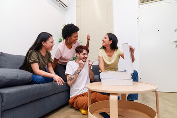 Fototapeta premium Joyful group of diverse young friends enjoying a pizza party at home. Excited man celebrates with cheerful women around a sofa.