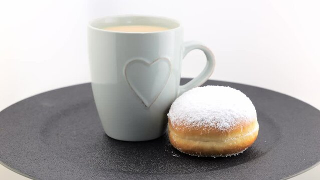 A cup of cappuccino and a freshly baked German doughnut with icing sugar, rotating on a black plate. Berlin donut (Berliner) or Krapfen.