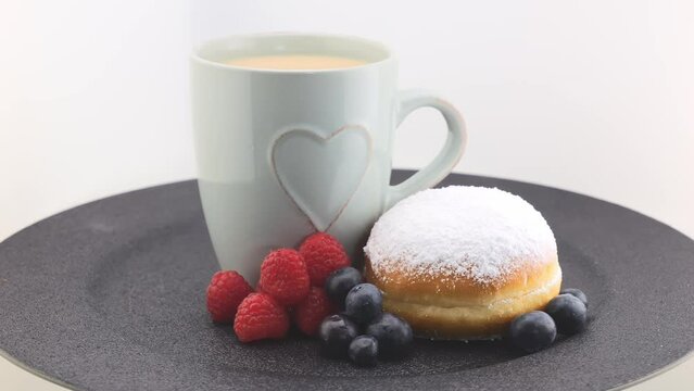 Cappuccino cup, German (or Italian) doughnut with icing sugar and berries (raspberries, blueberries) rotating on a black plate. Berlin donut (Berliner) or Krapfen. Italian donut Bomboloni.