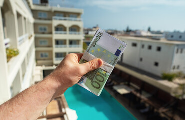 Man holds hundred-euro bills in front of a hotel with a swimming pool.