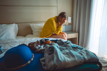 Flight cancellation. Upset woman sitting on bed with suitcases with clothes. - obrazy, fototapety, plakaty