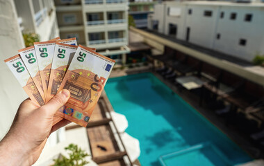 Man holds fifty-euro bills in front of a hotel with a swimming pool.
