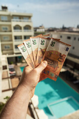 Man holds fifty-euro bills in front of a hotel with a swimming pool.