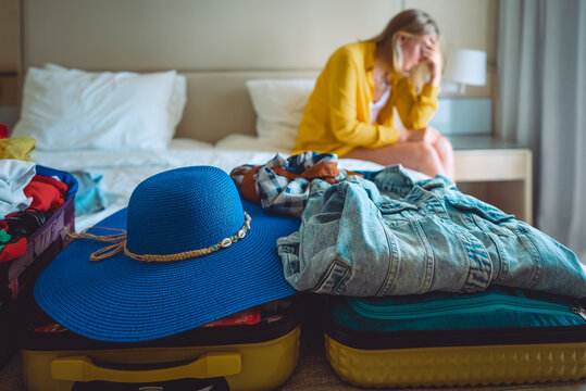 Flight cancellation. Upset woman sitting on bed with suitcases with clothes.