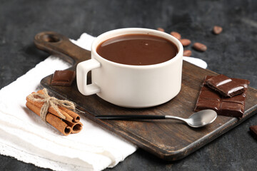 Wooden board with cup of tasty hot chocolate, cocoa beans and cinnamon on black grunge background