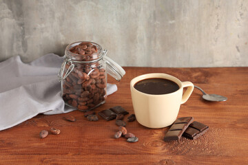 Cup of tasty hot chocolate and cocoa beans in jar on wooden table against grey grunge background