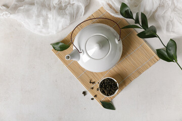 Bamboo mat with teapot and bowl of dry tea on light background