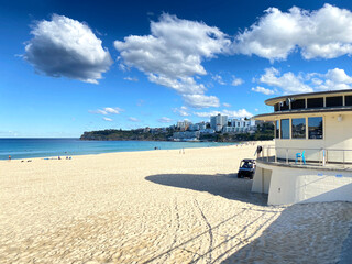 Obraz premium A day at the beach. Lifeguard hut on the sand to watch over people safety. Beach shore with sun and clouds. City’s buildings in the distance. 