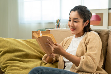 Asian beautiful woman sitting reading book on cozey couch sofa in living room with sun light...