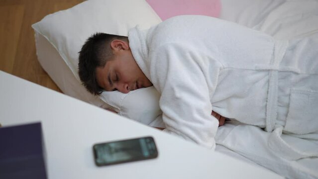 A young man in a white bathrobe continues to sleep, lying on a floor mattress in his bedroom, and does not respond to the sound of the alarm clock on his mobile phone lying on the nightstand