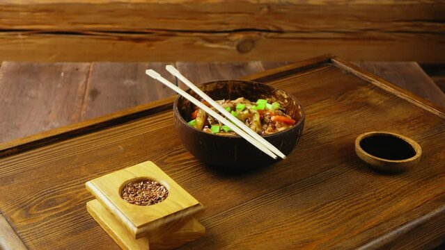 Soba with chicken and vegetables in a coconut shell plate next to the sticks on a wooden stand rotating in a circle. Close-up