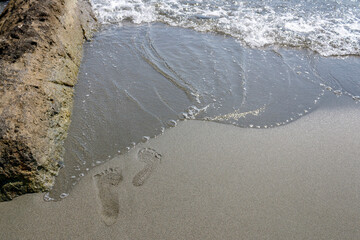 Sandy beach with two footprints close to sea wave and big rock