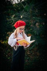 A little blonde girl in a red beret and suspenders is reading a book in nature