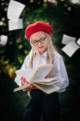 A little blonde girl in a red beret and suspenders is reading a book in nature