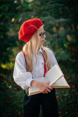 A little blonde girl in a red beret and suspenders stands with a yellow book in nature