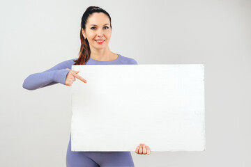 A friendly woman in a lilac fitness outfit smiles as she confidently holds a large, blank white sign, ideal for custom messages and advertising