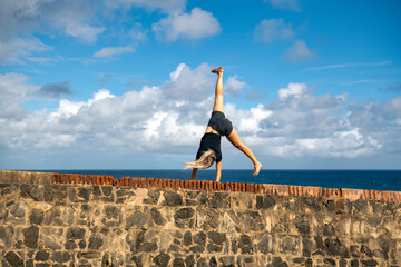 A young blonde woman does a cartwheel / hand stand on a stone wall along the sea.