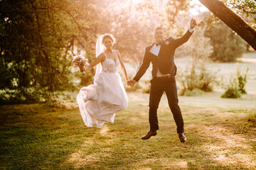 Valmiera, Latvia - July 7, 2023 - A joyful bride and groom jumping and holding hands in a sunlit...