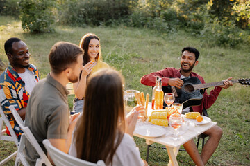 Meeting of multiracial group of friends playing guitar, singing, eating dinner and drinking wine during party in the forest