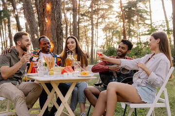 Meeting of multiracial group of friends playing guitar, singing, eating dinner and drinking wine during party in the forest