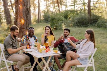 Meeting of multiracial group of friends playing guitar, singing, eating dinner and drinking wine during party in the forest