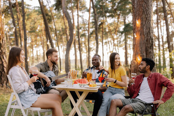 Meeting of multiracial group of friends playing guitar, singing, eating dinner and drinking wine during party in the forest