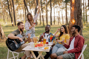Meeting of multiracial group of friends playing guitar, singing, eating dinner and drinking wine during party in the forest