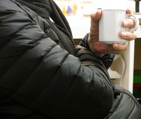 Sitzender Mann mit schwarzer Steppjacke und schwarzem Wollschal hält weißen Becher Kaffee in Hand...