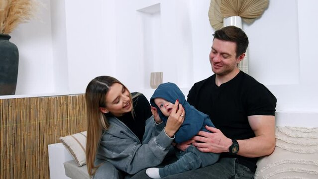 Cheerful young parents with infant child sit at home. Loving mom kisses her beloved baby.