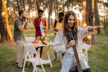 Beautiful hipster girl playing the guitar on the background of a dinner party outdoors of multiracial friends