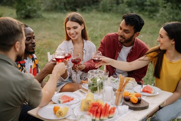 Multiracial group of happy friends sitting around table drinking wine talking at dinner party in evening summer forest