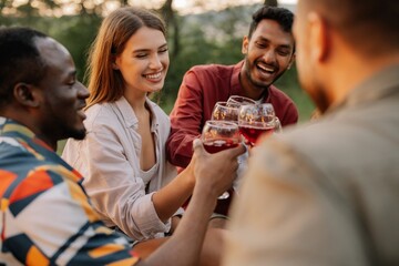 Multiracial group of happy friends sitting around table drinking wine talking at dinner party in evening summer forest
