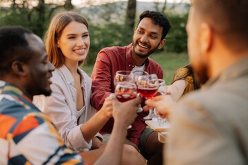 Multiracial group of happy friends sitting around table drinking wine talking at dinner party in evening summer forest