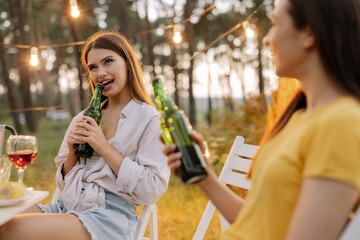 Funny hipster woman opens beer with teeth at party with friends in forest decorated with hanging lamps