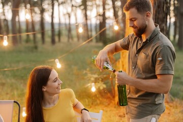 Bearded hipster man opens a beer at a party with friends in a forest decorated with hanging lamps