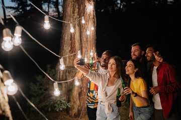 A group of multiracial friends having fun and drinking beer take selfies near hanging lamps at night