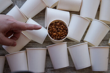 A finger points to a paper cup with coffee beans among empty cups. Fresh coffee beans
