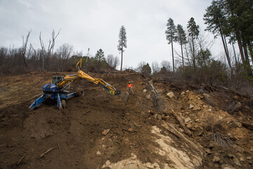 Erosion control/road work on steep hill above highway in Northern California after powerful atmospheric river storm.   Excavator and laborer can be seen working together.  