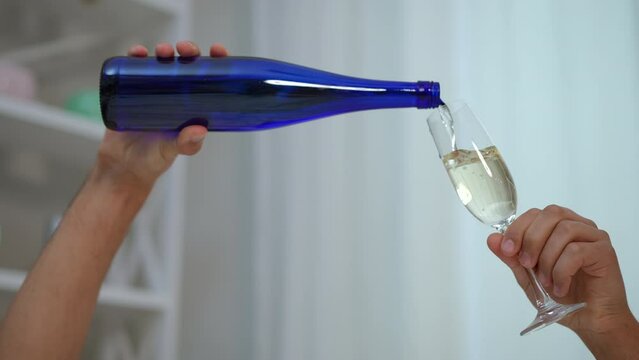 Slow motion. Close-up. Male hands pour white wine from a blue narrow bottle into a wine glass, against the backdrop of a home interior
