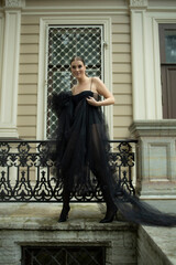 A 30-year-old girl in a sexy designer black dress and shoes poses in front of a house. Photo shoot, stylish look in a translucent dress. Fashion changes, trends, loneliness, widow, homewrecker