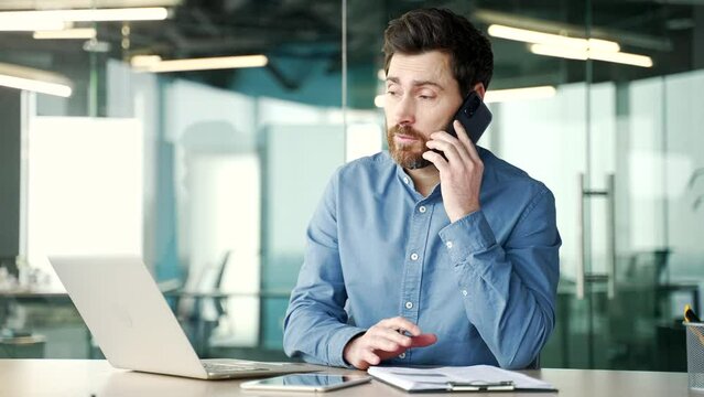 Busy manager talking on phone while sitting at workplace in modern office. Handsome bearded businessman communicates with a client, answers questions or conducts a conversation with a business partner