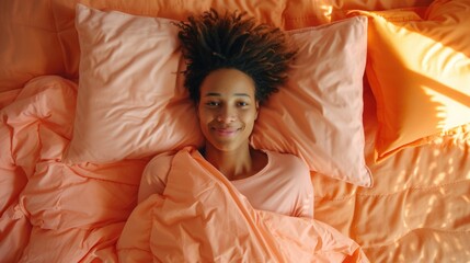An overhead view of a smiling woman lying in bed, covered with a peach fuzz color blanket