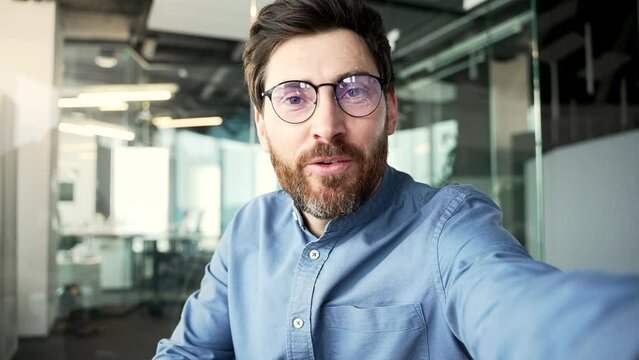 POV A smiling bearded businessman in glasses talking on a video call using a phone in a modern office. Handsome manager waving hand looking at camera. Online chat with a client or business partner