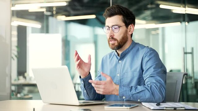Confident handsome businessman talking on a video call using a laptop computer sitting at workplace in office. Entrepreneur has a business meeting. The manager speaks remotely at an online conference