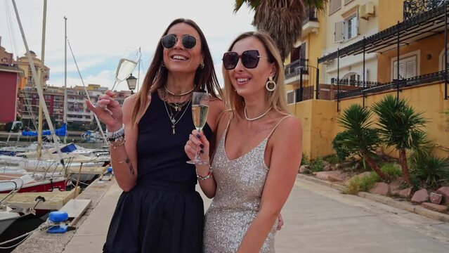 two women drink champagne and stand on a pier with yachts. sexy slender girlfriends hugging and smiling. Slow motion