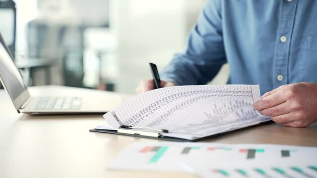 Close up of a man's hand signing documents at a desk at a workplace in a business office. Businessman in a blue shirt looks through the folder with documentation and signs the contract with a pen